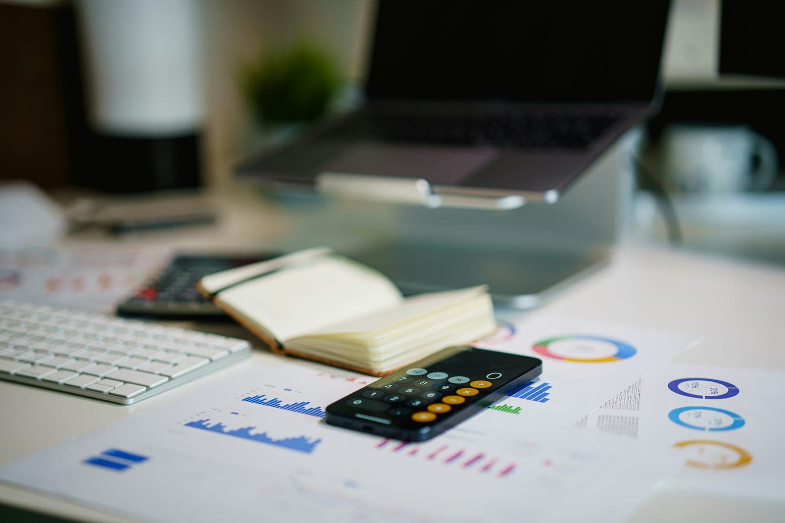 A phone calculator, an open notebook and printed charts on a desk in front of a laptop.